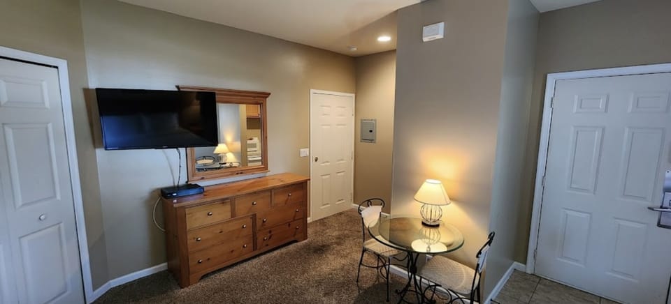 Bedroom sitting area with dresser storage, wall-mounted TV, small table with seating, and warm lighting.