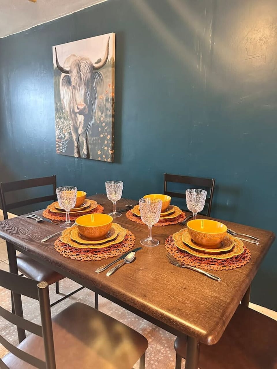 This dining area features a dark wood table elegantly set for four with orange accents against a backdrop of bold teal walls, enhanced by a striking Highland cow artwork. The setup creates a stylish and inviting space for memorable meals.