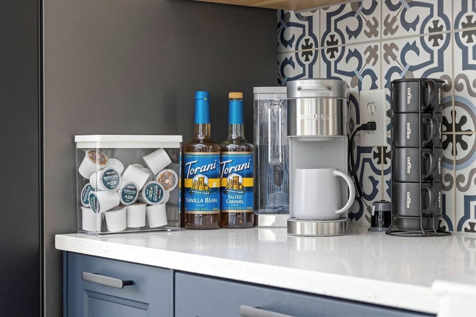 Kitchen counter corner with Keurig coffee maker, ground coffee K-cups, and syrup flavoring.
