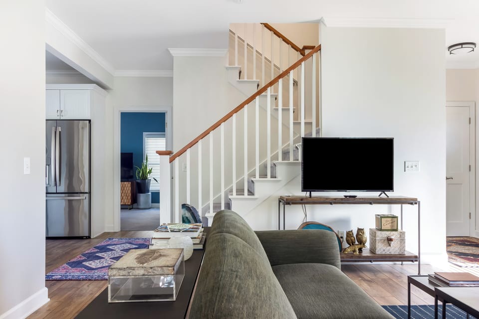 Modern living room with a cozy sofa, a TV on a stylish stand, and an inviting atmosphere. The space flows seamlessly into the dining area, with a staircase leading to the upper floors.







