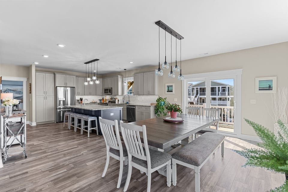 Bright open kitchen with gray cabinets, stainless appliances, white tile backsplash, pendant lights, large granite island with seating, wood-look floors, window view, and coastal décor in Oak Island.