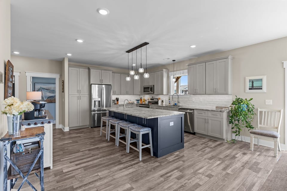 Bright open kitchen with gray cabinets, stainless appliances, white tile backsplash, pendant lights, large granite island with seating, wood-look floors, window view, and coastal décor in Oak Island.