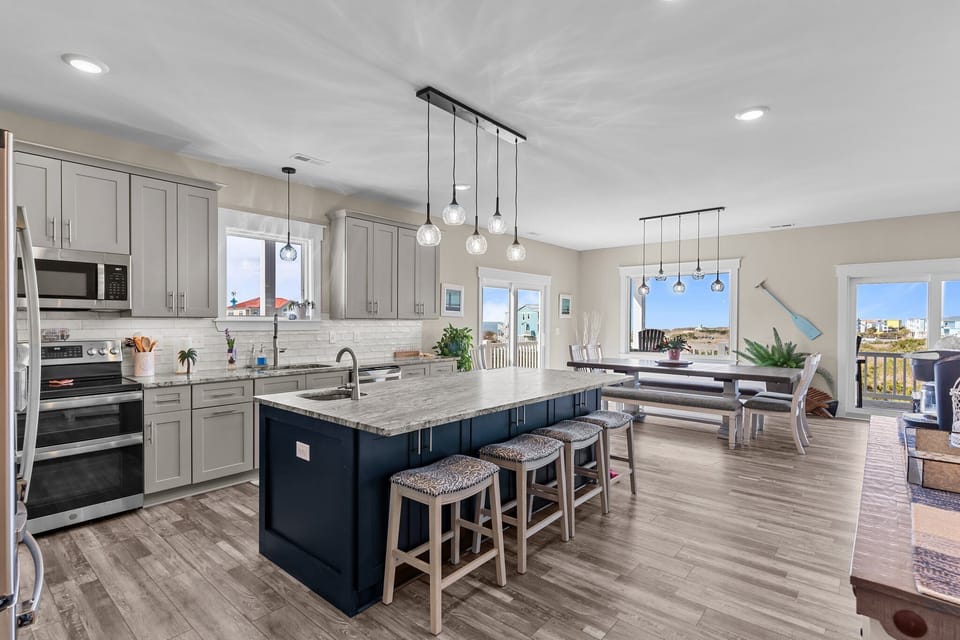 Bright open kitchen with gray cabinets, stainless appliances, white tile backsplash, pendant lights, large granite island with seating, wood-look floors, window view, and coastal décor in Oak Island.