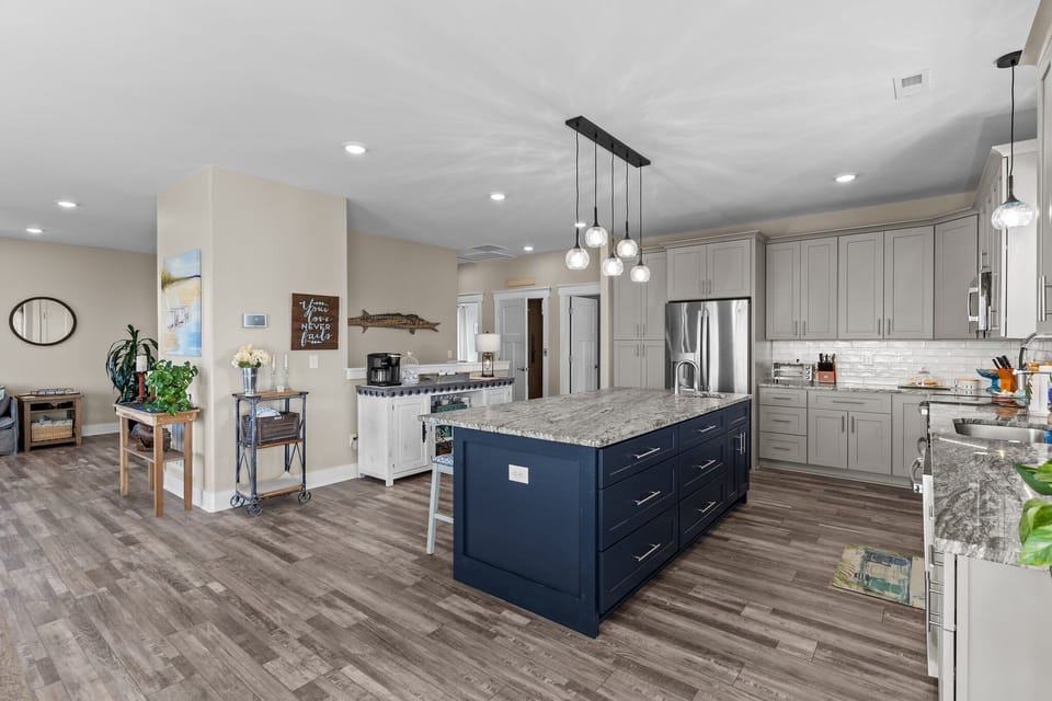 Bright open kitchen with gray cabinets, stainless appliances, white tile backsplash, pendant lights, large granite island with seating, wood-look floors, window view, and coastal décor in Oak Island.