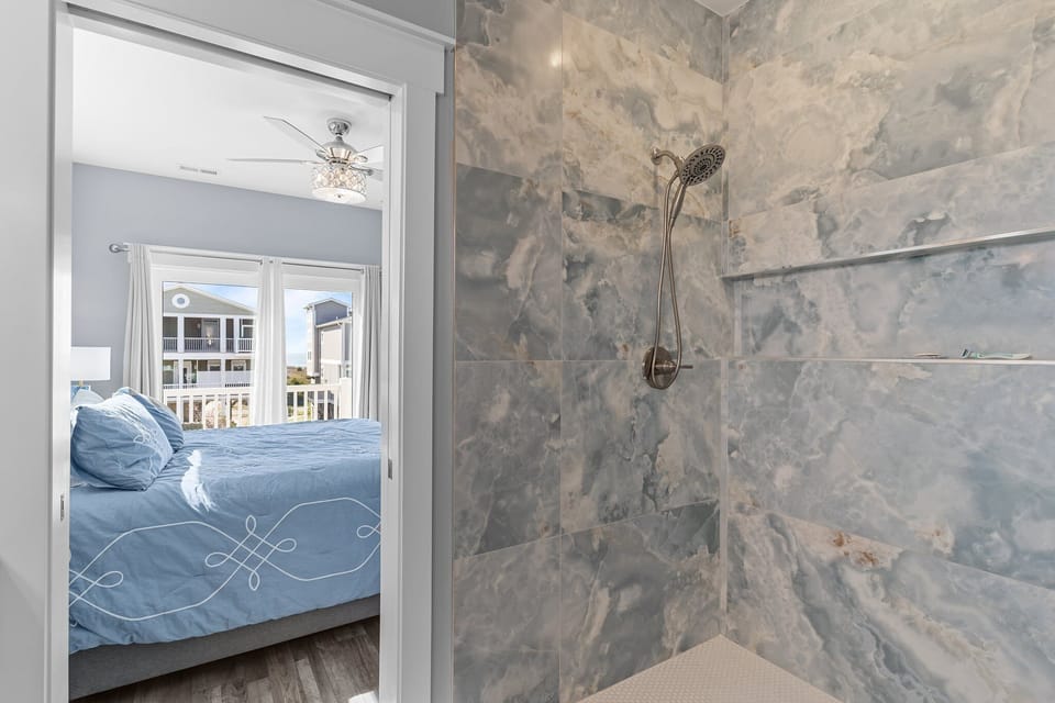 Modern walk-in shower with large blue marble-look tile walls, built-in shelf, brushed nickel showerhead, and doorway view into king bedroom with balcony access in this Oak Island beach home.