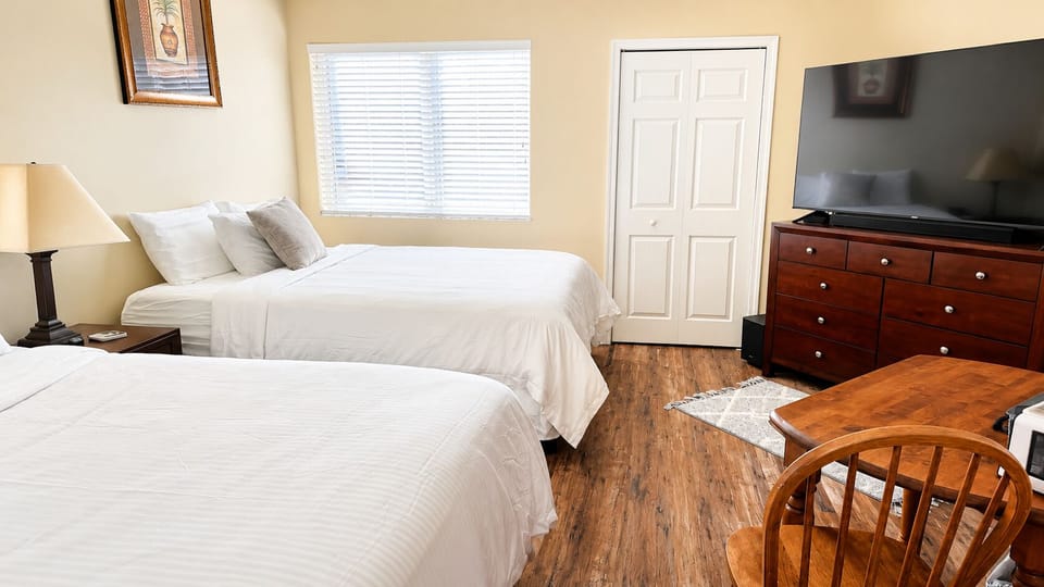 Bright bedroom featuring two beds, dresser storage, TV, wood-look flooring, and a cozy table with seating.