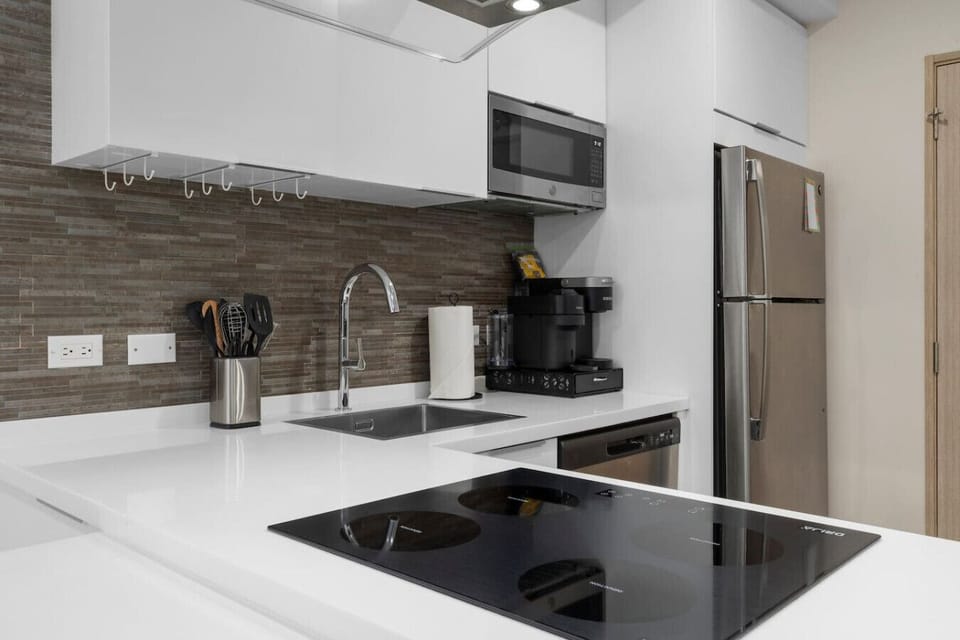 A modern kitchen with a clean design. The kitchen features white cabinets, a black stovetop, and stainless steel appliances. The backsplash is a textured brown and gray tile.