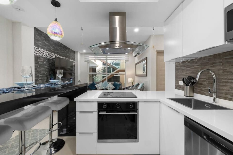 A modern kitchen with a black island and white barstools. White cabinets and stainless steel appliances, including a microwave, dishwasher, and refrigerator, are visible. The backsplash is a gray textured tile.