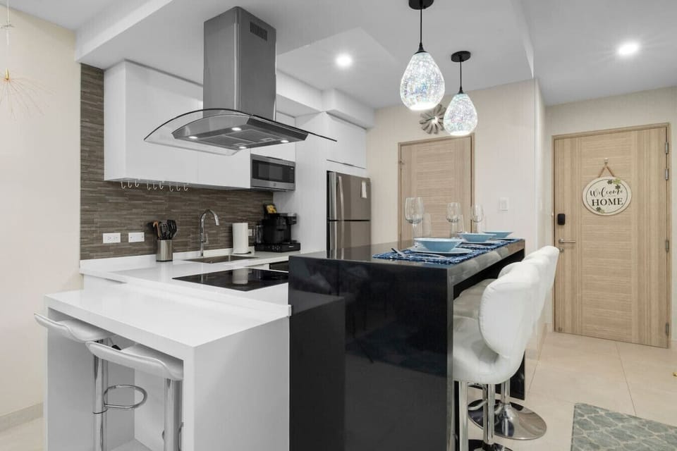 Modern kitchen with a black island and white countertops. It features white cabinets, stainless steel appliances, and a gray backsplash. Pendant lights hang above the island, and a `Welcome Home` sign is on a door.