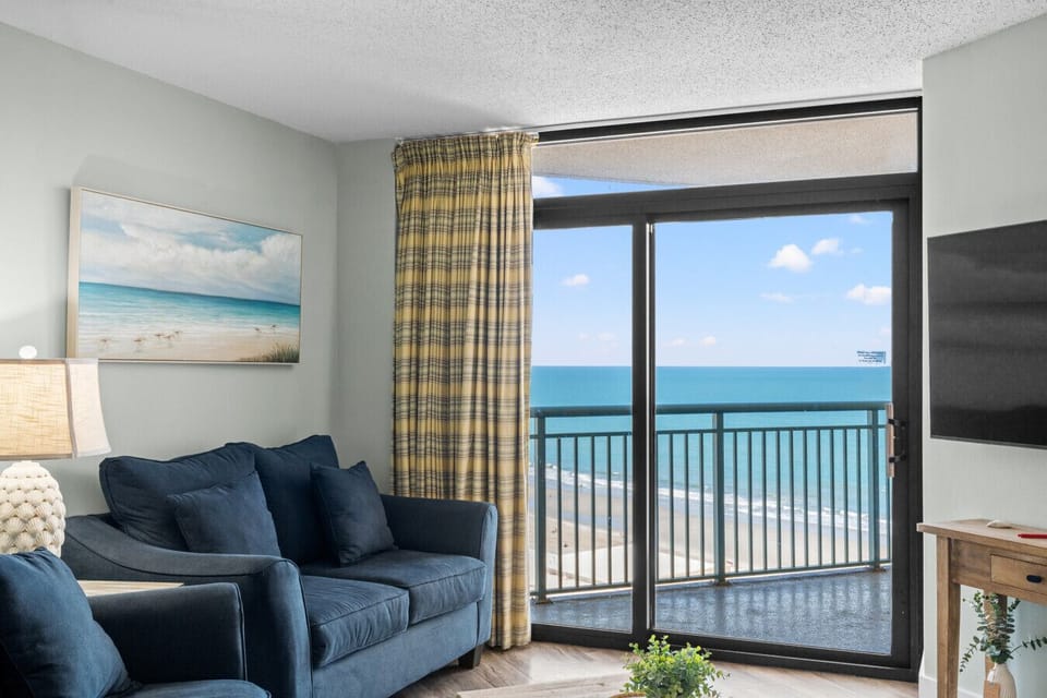 Spacious oceanfront living room with oversized sofa and loveseat, smart TV, and direct balcony access. Floor-to-ceiling windows flood the space with natural light and showcase stunning Myrtle Beach coastline views.