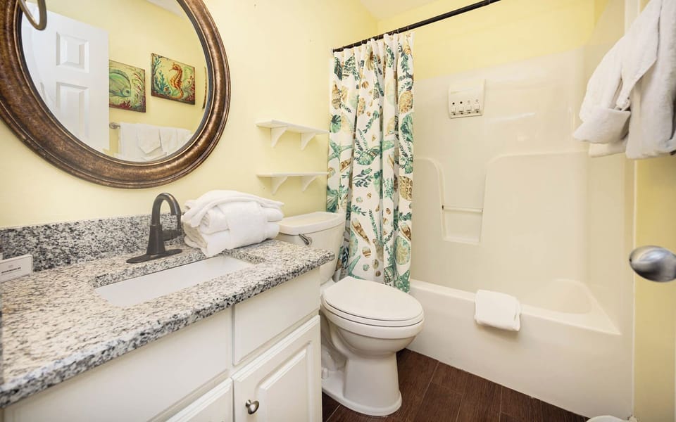 The bathroom features a granite countertop, a round mirror, and a colorful shower curtain, for a relaxing experience