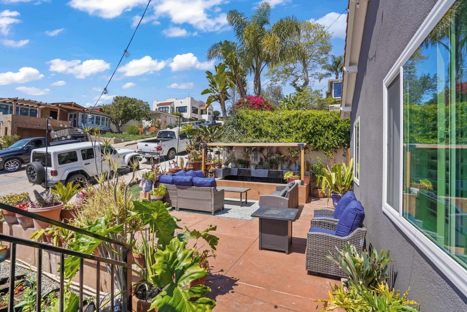 This inviting outdoor patio features comfortable wicker seating with blue cushions and a stylish gray set. A pergola offers shade, while numerous potted plants add a touch of nature. Enjoy the sunny sky and neighborhood views from this relaxing space