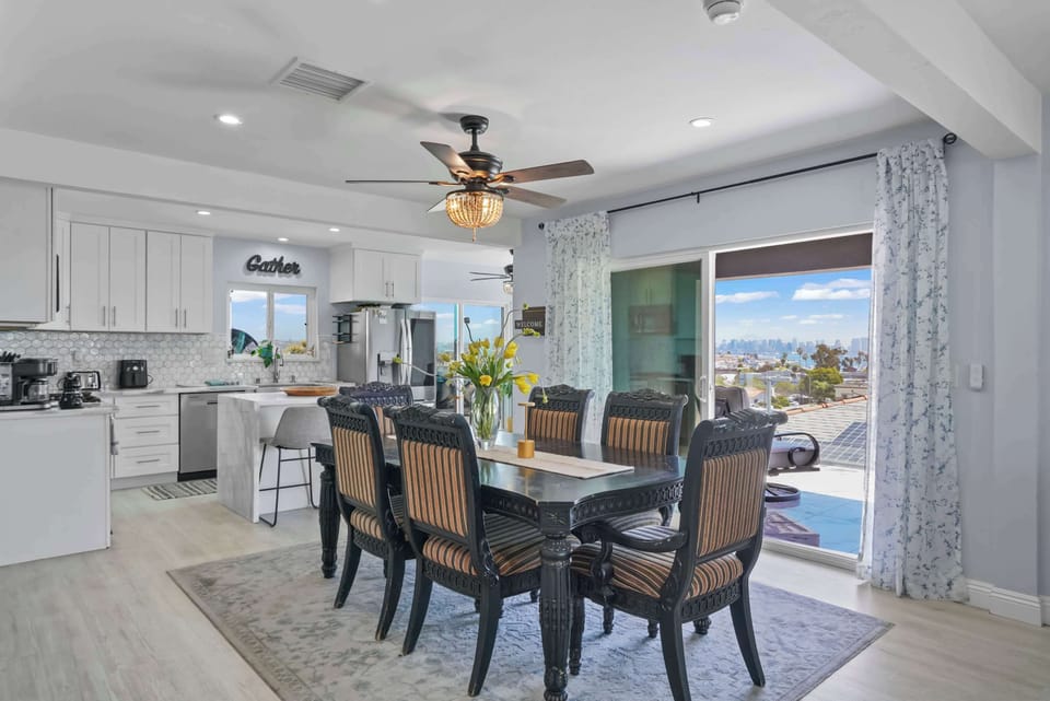 Open dining area with a dark wood table and striped chairs on a gray rug. Yellow flowers add a vibrant touch. The space flows into a modern white kitchen and offers views of an outdoor pool and cityscape through sliding glass doors.