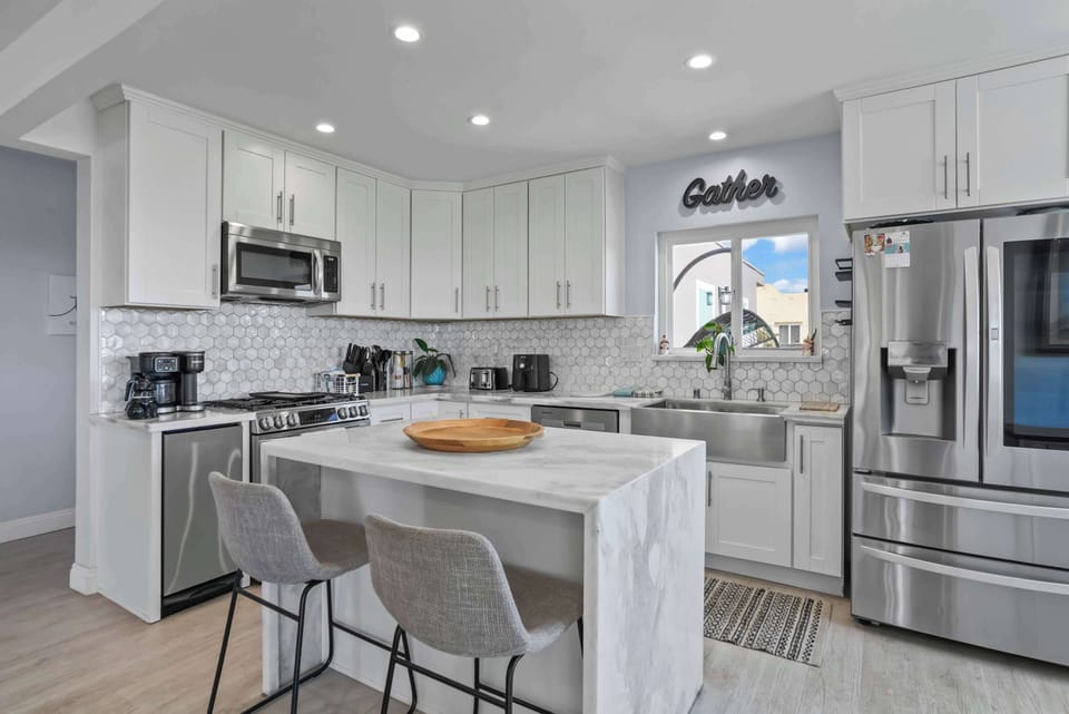 This bright, modern kitchen features sleek white cabinetry, stainless steel appliances, and a stylish marble-top island with gray bar stools. Natural light fills the space, highlighting the white tiled backsplash and the "Gather" sign above the sink.