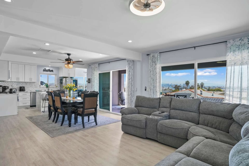 Bright, open-concept living space with scenic views from large windows. Features a comfortable gray sectional sofa, a dining area with a dark table and chairs, and a modern white kitchen visible in the background. A ceiling fan and recessed lighting.