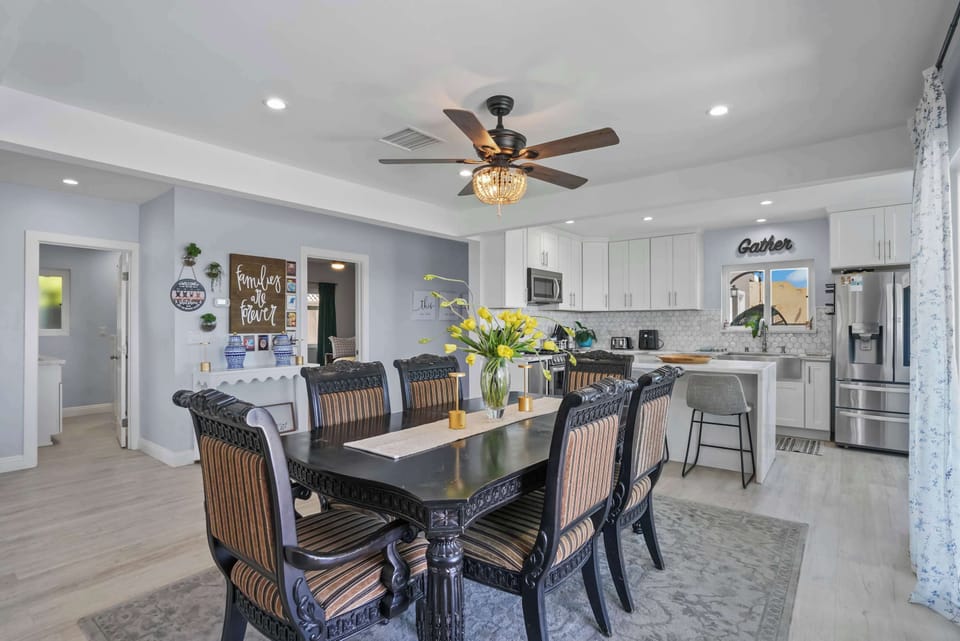 This bright and airy dining area features a dark wood table with striped chairs, perfect for gatherings. Yellow flowers add a pop of color. The open-plan layout flows into a modern kitchen with white cabinets and stainless steel appliances, illuminat