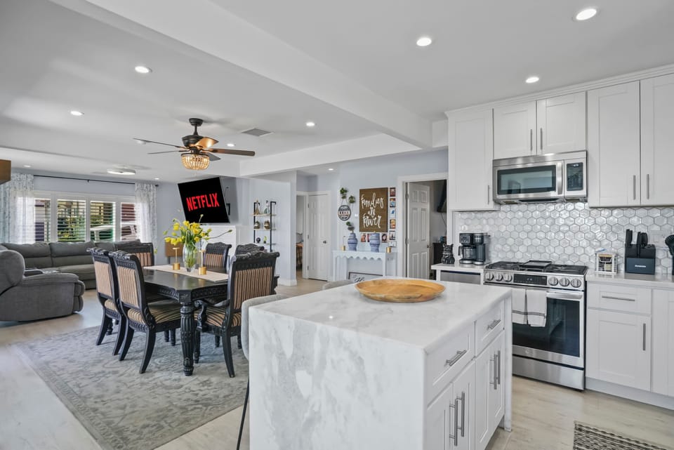 This open-concept living space seamlessly blends a modern white kitchen with a dining area featuring a dark table and chairs. Beyond, a comfortable living room with a large TV  invites relaxation. Recessed lighting illuminates the entire area.