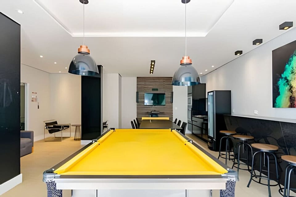 Sala de jogos com mesa de sinuca, mesa 6 cadeiras, balcão 4 cadeiras, televisão, sofá e geladeira.