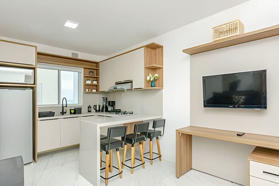 Cozinha moderna e completa com geladeira, pia, cooktop 4 bocas, exaustor, pia, armários, micro-ondas, chaleira elétrica, liquidificador, cafeteira, balcão 3 lugares com utensílios de café da tarde e televisão.