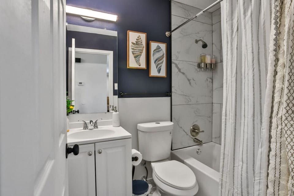 Full bath with marble-tile tub/shower, navy accent wall, white vanity, and coastal art — stylish and fully stocked