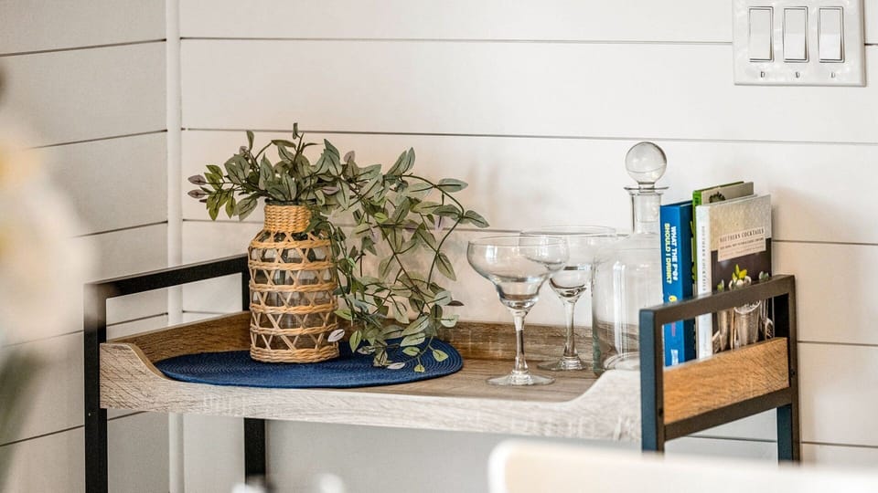 Bright and coastal dining area with a table set for eight, offering views of the charming homes and condos along the street. Features include dinner service for eight, a bar cart ready for cocktail hour, and access to the kitchen for seamless entertainment