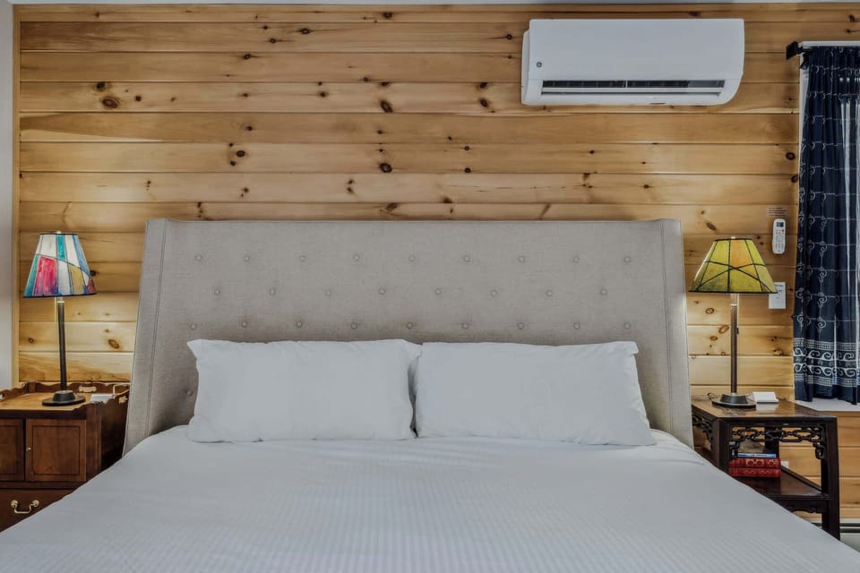King bed with tufted headboard, stained glass lamps, knotty pine accent wall, and mini-split AC