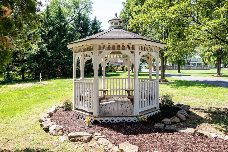 A white wooden gazebo surrounded by lush greenery and a stone-bordered flower bed.