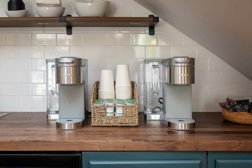 Two stainless steel coffee makers with cups and pods on a wooden countertop under a wooden shelf.