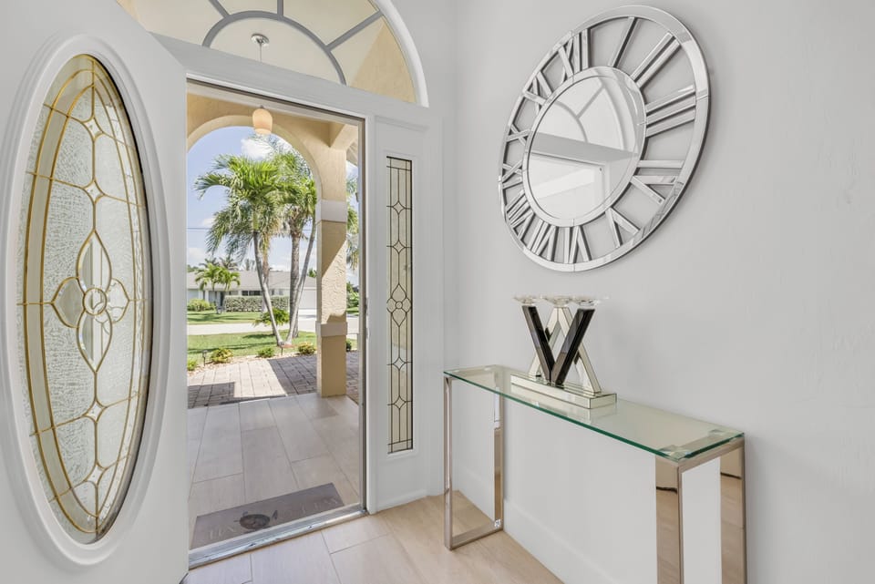 - Step inside through frosted glass doors into a sunlit entry with tropical views ahead
- Bold silver wall clock and sleek console table offer modern coastal elegance
- Stunning entrance sets the tone for your vacation retreat