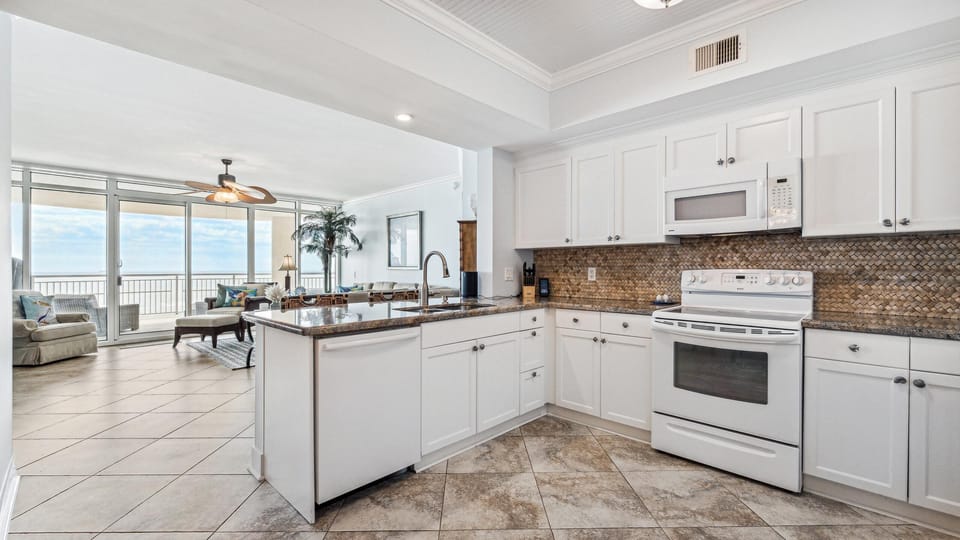 Bright, coastal kitchen with white cabinetry, granite countertops, and mosaic tile backsplash flows effortlessly into an airy living area with floor-to-ceiling windows and stunning Gulf views.