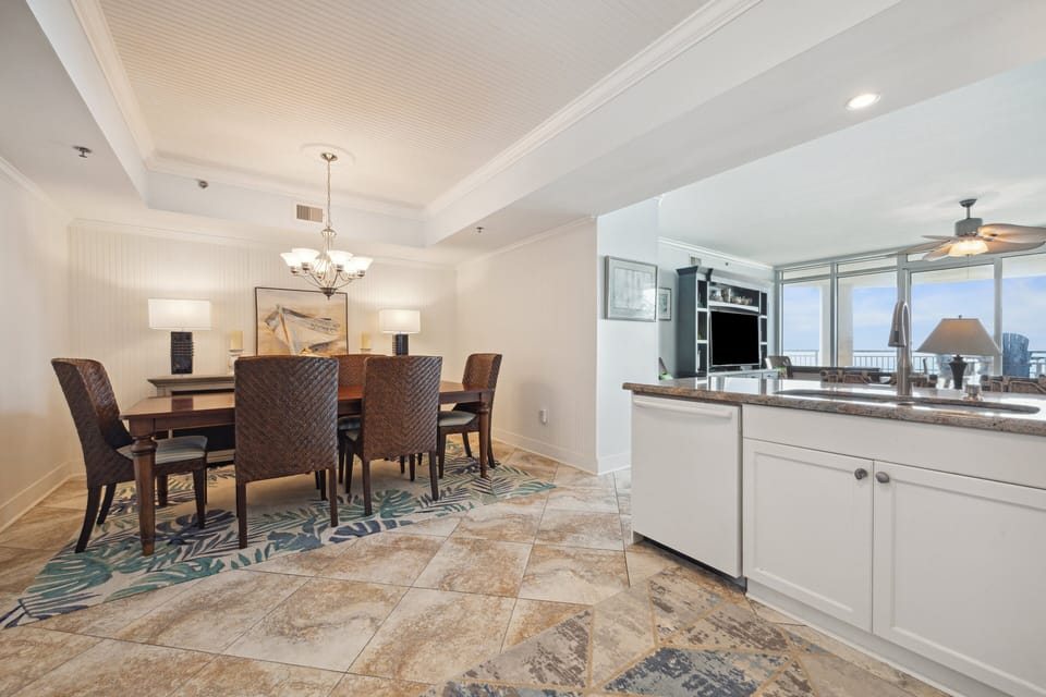 Elegant dining area with seating for six, accented by tropical woven chairs and ambient lighting, seamlessly connected to an open-concept kitchen with granite countertops and ocean views.