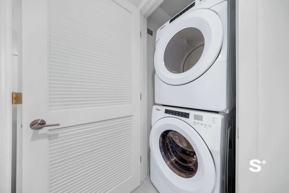 A compact apartment features an enclosed laundry closet with stacked, front-loading washer and dryer units neatly hidden behind a white louvered door.