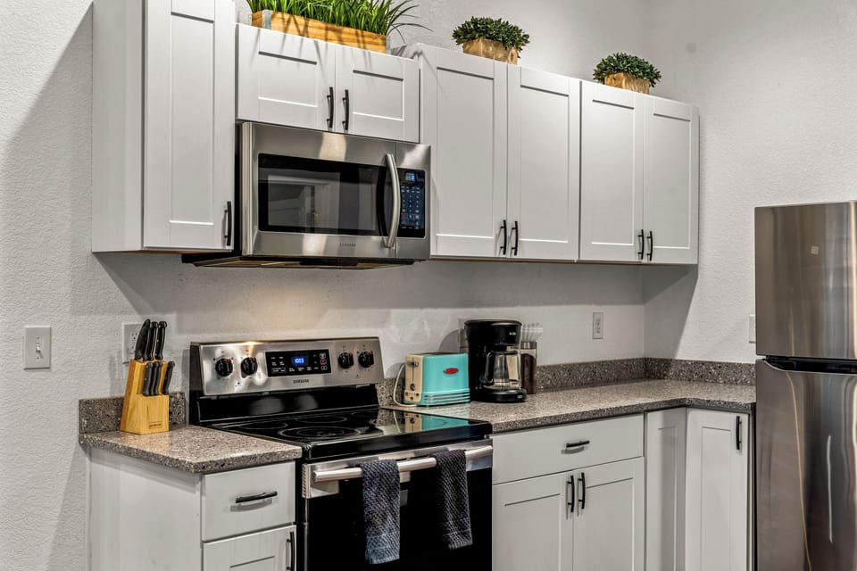 - Stainless steel appliances paired with crisp white cabinetry and modern decor
- Includes coffee maker, microwave, toaster, and fully stocked drawers
- Decorative greenery adds freshness to this clean and functional space