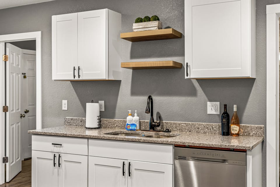- Granite countertops, sleek black faucet, and floating wood shelves add modern flair
- Stocked with hand soap, paper towels, and essentials for your convenience
- Stainless steel dishwasher completes this clean and functional space
