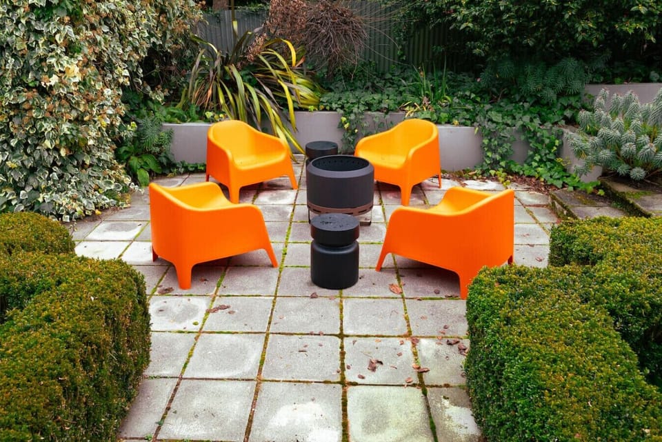 Outdoor patio featuring four burnt orange lounge chairs, a cosy firepit, handy coffee tables, all nestled in lush, vibrant gardens.