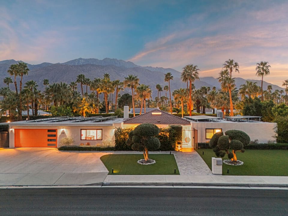 Twilight exterior showing the home glowing against mountain views & palm-lined landscaping.