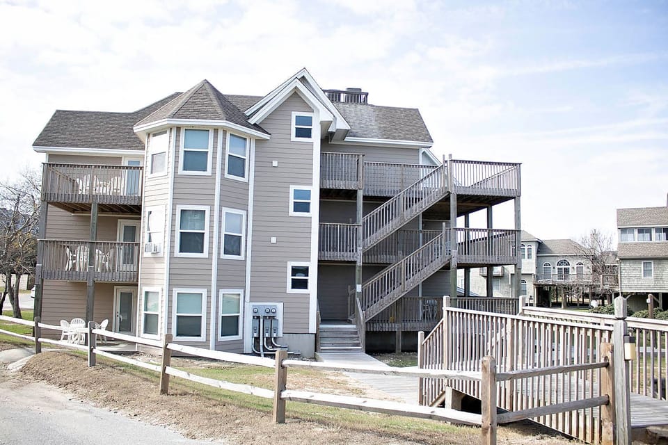 A multi-story apartment building with a tan exterior, wooden balconies, and a set of stairs, situated near a grassy area and wooden fence under a partly cloudy sky.