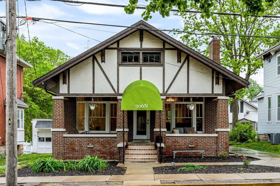 A straight-on arrival view showcases the historic brick bungalow, green 306N awning, covered porch, and mature landscaping.