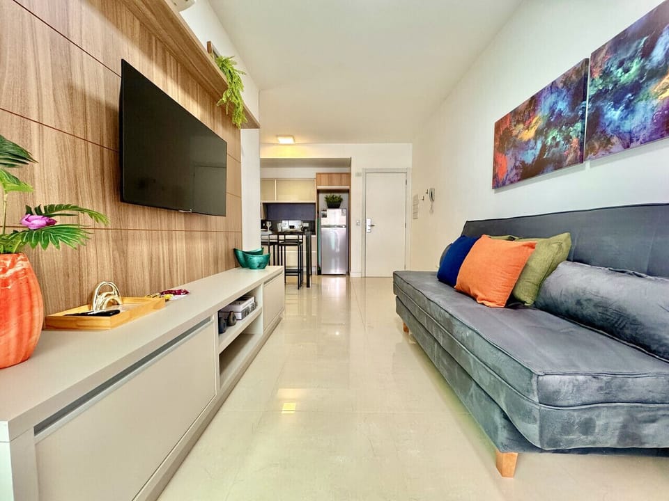 Corredor com sofa cinza, Smart TV na parede e acesso a cozinha