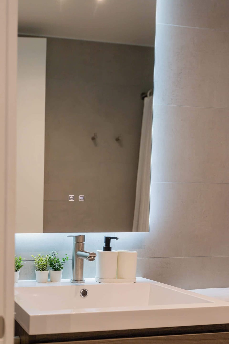 Experience modern elegance in this stylish bathroom, featuring a sleek sink, backlit mirror, and decorative plants for a refreshing touch.