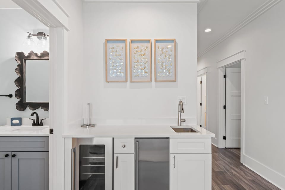 Your private wet bar offers everything you need to unwind—a sleek sink, wine cooler, and mini fridge, all framed by elegant coastal decor and warm lighting.