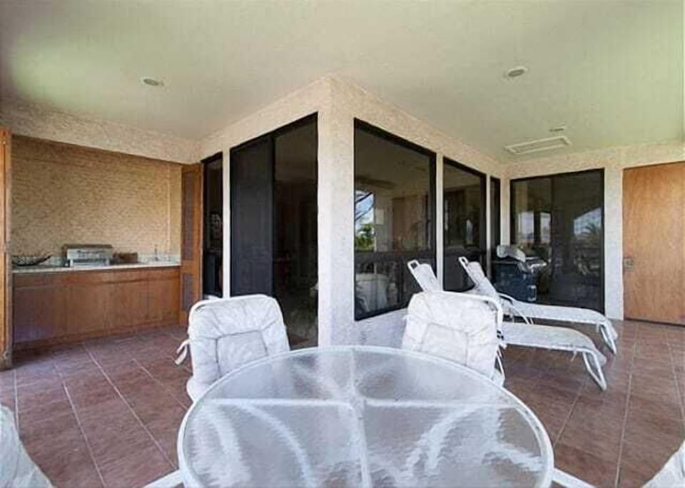 Covered lanai with cushioned loungers, glass dining set, and a built in grill station, perfect for al fresco meals.