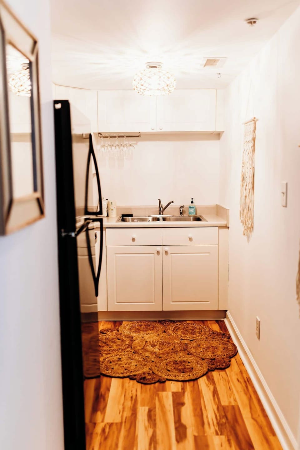 This boho-chic kitchen features full-sized appliances, including a black fridge and stove, plus a double sink. White cabinets, a jute rug, and a wine glass rack add warmth and style—perfect for cooking, sipping, or settling in with ease.
