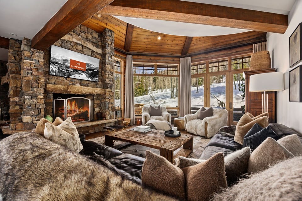 View of a luxury living room with ski slopes visible through a wall of windows. The couch is large and overstuffed with many pillows. there are two faux fur covered couches, a large fireplace and a TV