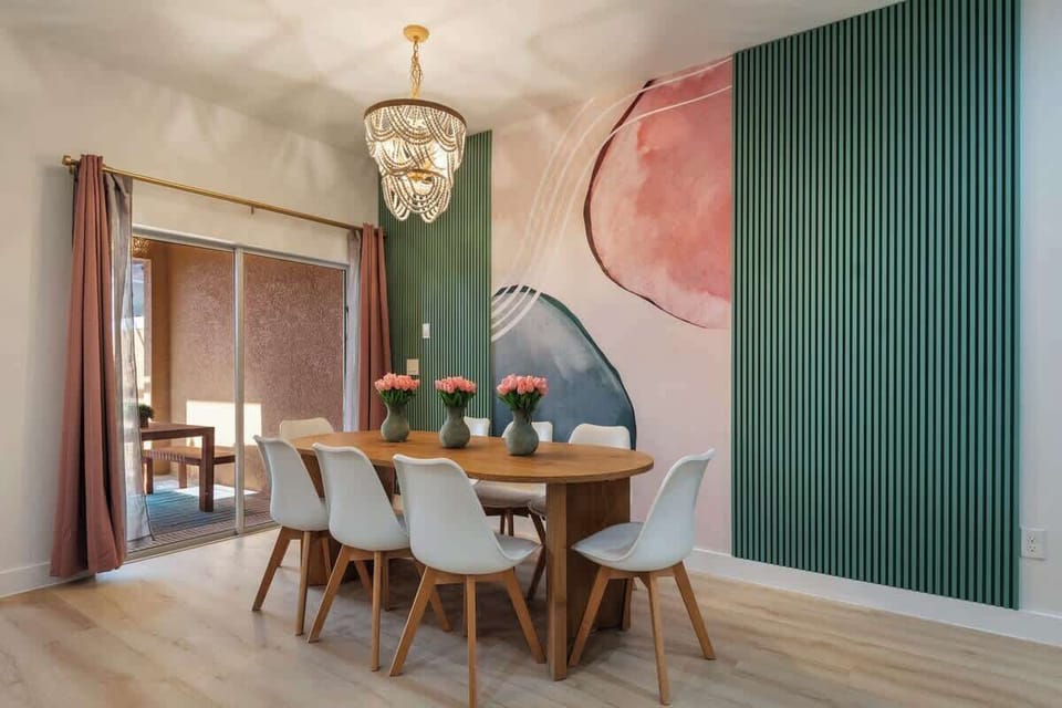 Stylish dining area with chic chandelier, vibrant mural, and elegant wooden table. Features modern white chairs and sliding glass doors for natural light. Perfect for gatherings or relaxing. Experience comfort and style—reserve your stay today!