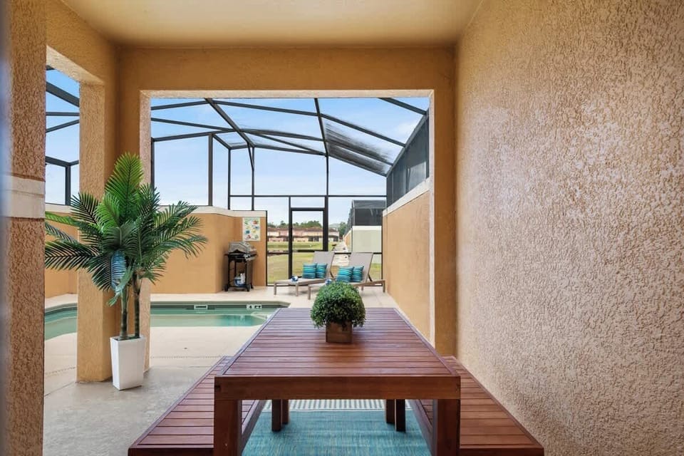 Screened outdoor area with a wooden table and benches, adorned with potted plants. Overlooks a pool with lounge chairs and a grill, surrounded by warm-toned walls. Offers a cozy, private space for dining and relaxation.
