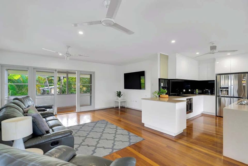 A bright and airy living area with sleek timber floors, plush lounge seating, and seamless flow to the modern kitchen and leafy outdoor views.