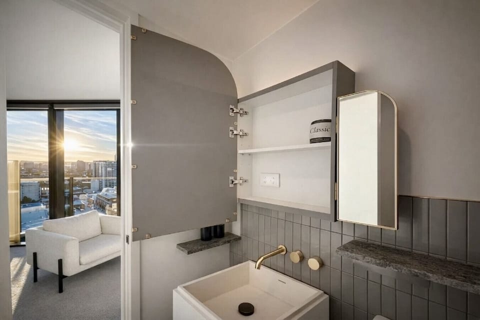 Designer ensuite with brass tapware, vertical grey tiling, and a stone-topped vanity. Open mirrored cabinet offers practical storage, while the adjoining bedroom reveals a sunset city view through the doorway.