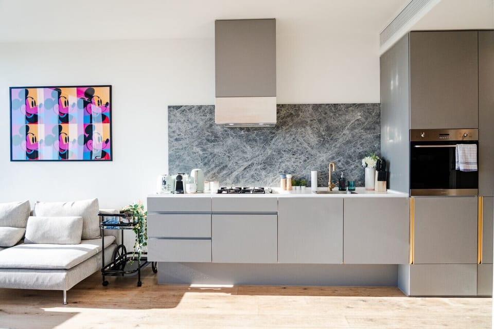 Sleek kitchen with grey handleless cabinetry, a marble-look stone splashback, gold tap, and built-in oven, opening to the living area. A vibrant pop-art canvas and warm timber floors add personality to the space.