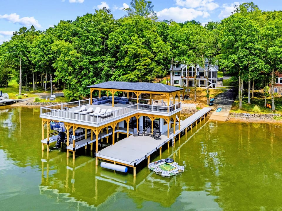 Aerial - The boathouse from the water, upper deck loungers and covered slips below, the home set back in the trees.